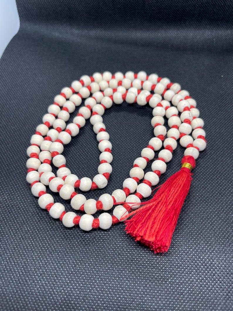 May include: A strand of round, off-white beads strung on a vibrant red cord, arranged in a circular loop. The necklace features a prominent red tassel at the bottom, secured with a small gold-coloured band, creating a continuous, knotted design.