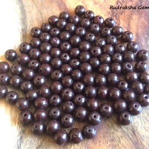 May include: A pile of dark brown round beads, likely made of wood. The beads are smooth and shiny, with a slight reddish hue. Rudraksha Gems is written in white text on a brown background.