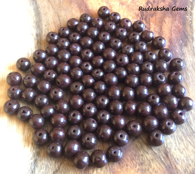 May include: A pile of dark brown round beads, likely made of wood. The beads are smooth and shiny, with a slight reddish hue. Rudraksha Gems is written in white text on a brown background.