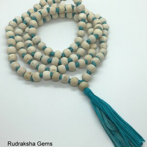 May include: A long strand of light beige wooden beads strung on a vibrant turquoise cord, arranged in a circular pattern. The necklace features a matching turquoise tassel at the bottom and includes the text Rudraksha Gems in the corner.