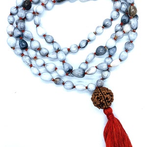 May include: A grey and white Rudraksha mala with a red tassel and a brown Rudraksha bead. The mala is made of seeds and is used for meditation and prayer.