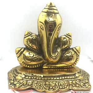 Puede incluir: Una estatua dorada de Ganesh con detalles intrincados. La estatua presenta una cabeza grande, orejas prominentes y un tronco. Tres gemas rojas adornan la frente. La base est&aacute; decorada con patrones ornamentados. La estatua es de metal.