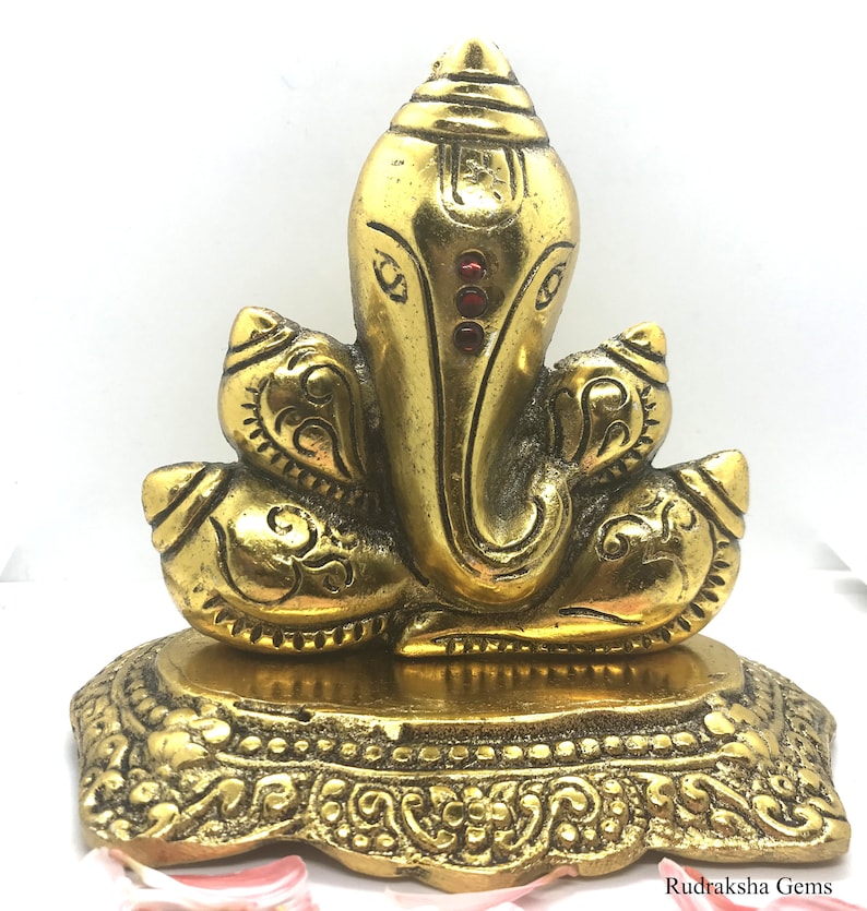 Puede incluir: Una estatua dorada de Ganesh con detalles intrincados. La estatua presenta una cabeza grande, orejas prominentes y un tronco. Tres gemas rojas adornan la frente. La base est&aacute; decorada con patrones ornamentados. La estatua es de metal.