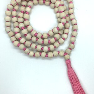 May include: A long strand of light beige wooden beads strung on a bright pink cord, arranged in a circular coil. The necklace features a matching pink tassel at the end. The text Rudraksha Gems is printed in black in the bottom left corner.