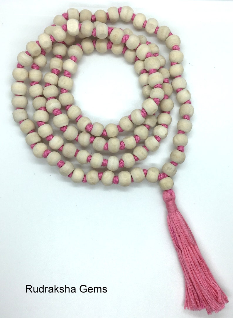 May include: A long strand of light beige wooden beads strung on a bright pink cord, arranged in a circular coil. The necklace features a matching pink tassel at the end. The text Rudraksha Gems is printed in black in the bottom left corner.