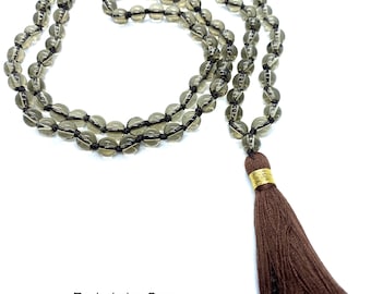 May include: A brown mala necklace with a tassel made of dark brown fabric. The necklace is made of round, translucent, smoky quartz beads.