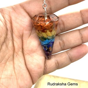 Op de afbeelding: Een zeven chakra kristallen slinger met een zilveren ketting. De slinger is gemaakt van helder hars met ingebedde kristallen in de kleuren van de zeven chakra's: rood, oranje, geel, groen, blauw, indigo en violet.