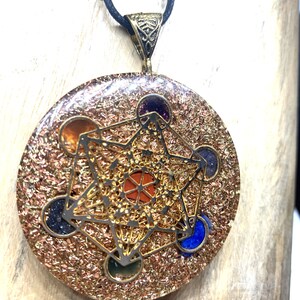 May include: A round pendant with a Metatron's Cube design, featuring a gold-colored filigree overlay and embedded gemstones in various colors. The pendant hangs from a black cord. The background is a light-colored wooden surface. The text "Rudraksha Gems" is at the bottom.