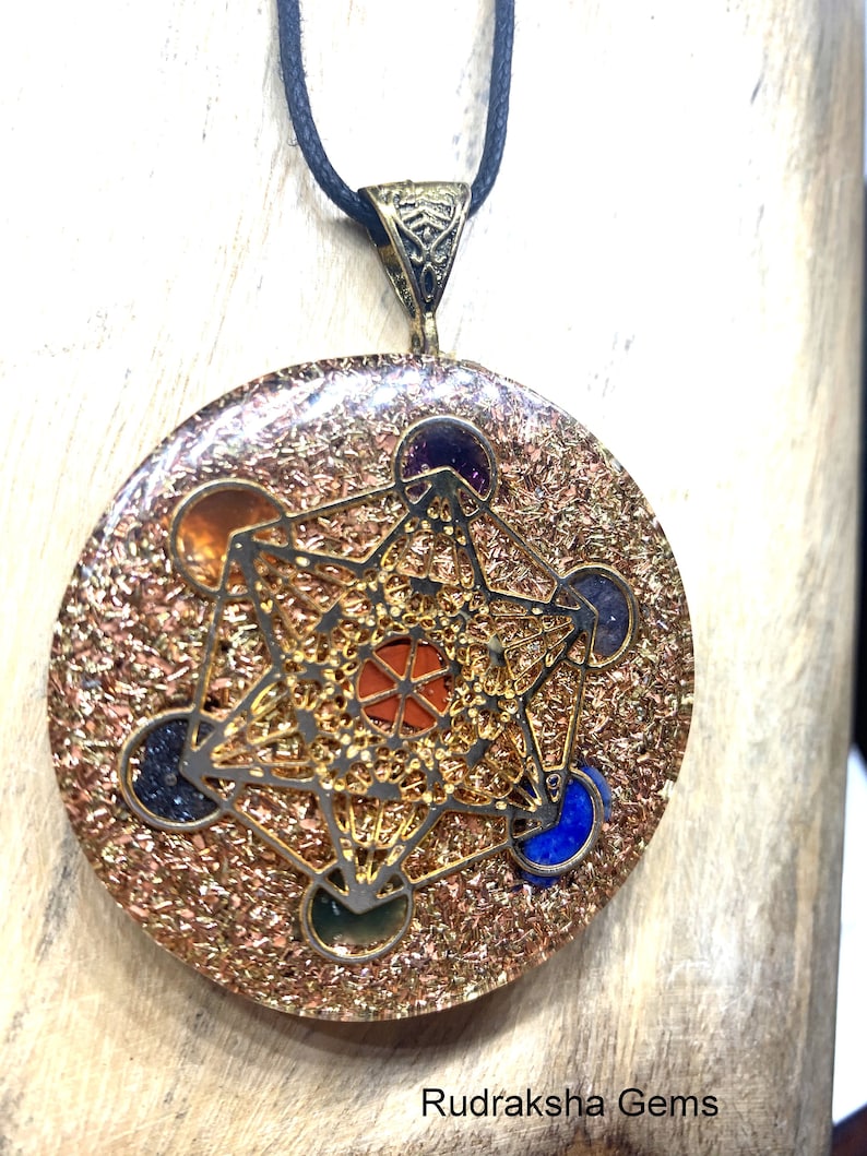 May include: A round pendant with a Metatron's Cube design, featuring a gold-colored filigree overlay and embedded gemstones in various colors. The pendant hangs from a black cord. The background is a light-colored wooden surface. The text "Rudraksha Gems" is at the bottom.