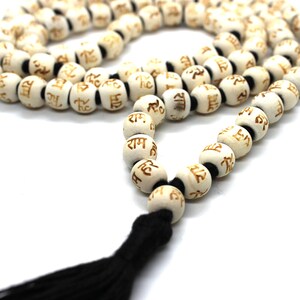 K&ouml;nnte beinhalten: Wei&szlig;e Holz-Mala-Perlen mit einem schwarzen Quasten. Jede Perle ist mit dem Wort "Ram" in goldenen Buchstaben graviert.