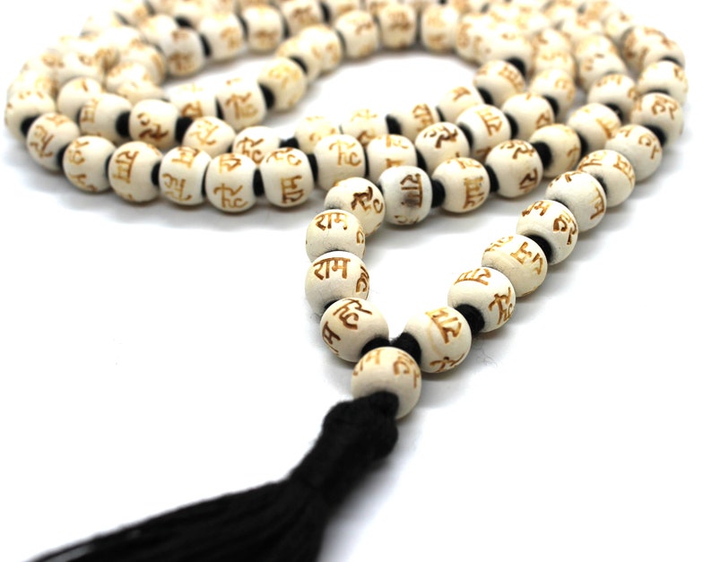 K&ouml;nnte beinhalten: Wei&szlig;e Holz-Mala-Perlen mit einem schwarzen Quasten. Jede Perle ist mit dem Wort "Ram" in goldenen Buchstaben graviert.