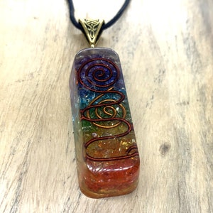 Peut inclure: Un pendentif en cristal arc-en-ciel avec un motif de fil de cuivre, suspendu à un cordon noir. Le pendentif est un prisme rectangulaire avec un sommet et une base arrondis. Le cristal est composé de différentes couleurs, dont le rouge, l'orange, le jaune, le vert, le bleu, l'indigo et le violet. Le motif de fil de cuivre est une spirale qui s'enroule autour du cristal. Le pendentif est suspendu à un cordon noir avec un fermoir doré. Le pendentif est sur une surface en bois. Rudraksha Gems.