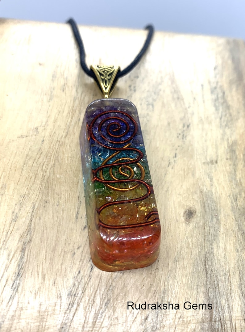 May include: A rainbow colored crystal pendant with a copper wire design, hanging from a black cord. The pendant is a rectangular prism with a rounded top and bottom. The crystal is layered with different colors, including red, orange, yellow, green, blue, indigo, and violet. The copper wire design is a spiral that wraps around the crystal. The pendant is hanging from a black cord with a gold-colored clasp. The pendant is on a wooden surface. Rudraksha Gems.