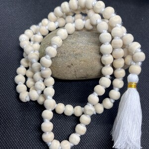May include: A strand of light-coloured, natural wood beads strung on a white cord, featuring a white fabric tassel at one end secured with a thin gold-coloured metallic band. The beads are round, uniform in size, and arranged in a circular loop.