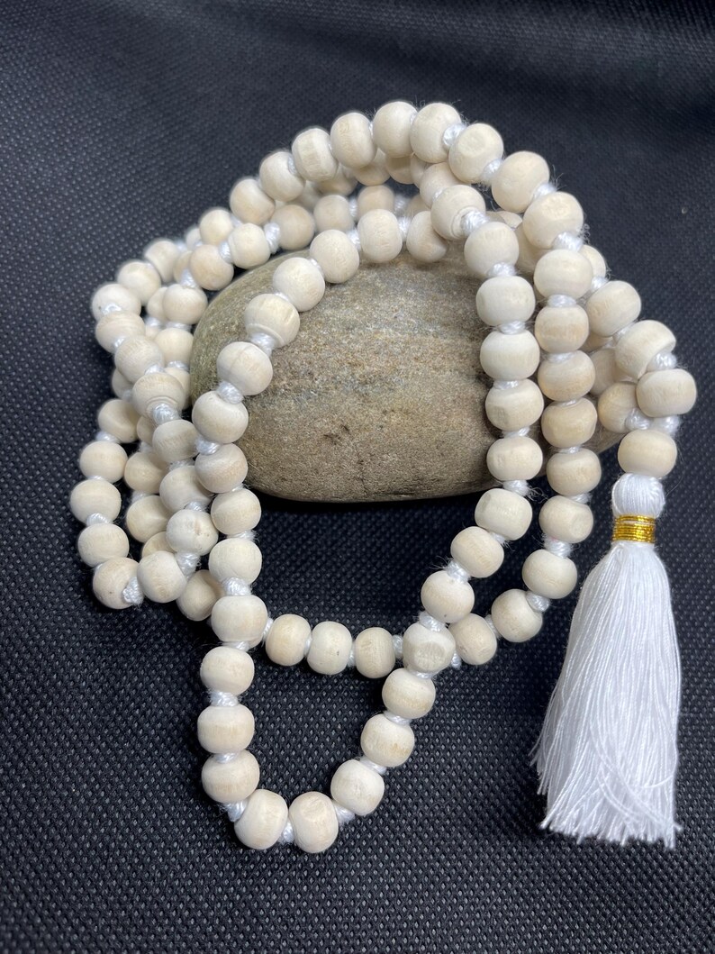 May include: A strand of light-coloured, natural wood beads strung on a white cord, featuring a white fabric tassel at one end secured with a thin gold-coloured metallic band. The beads are round, uniform in size, and arranged in a circular loop.