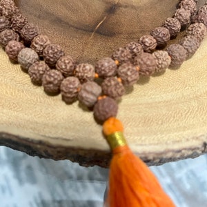 Peut inclure: Un collier de perles de mala en bois brun avec un pompon orange. Les perles sont en bois naturel et sont enfilées sur un cordon fin. Le pompon est en tissu orange doux.
