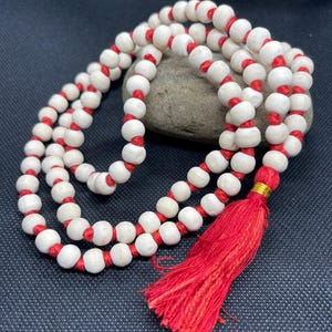May include: A strand of round, off-white wooden beads strung on a vibrant red cord. The necklace features a prominent red tassel at the end, secured with a small gold-coloured metallic band, creating a continuous loop of beads with a knotted design.