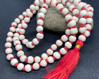 May include: A strand of round, off-white wooden beads strung on a vibrant red cord. The necklace features a prominent red tassel at the end, secured with a small gold-colored metallic band, creating a continuous loop of beads with a knotted design.