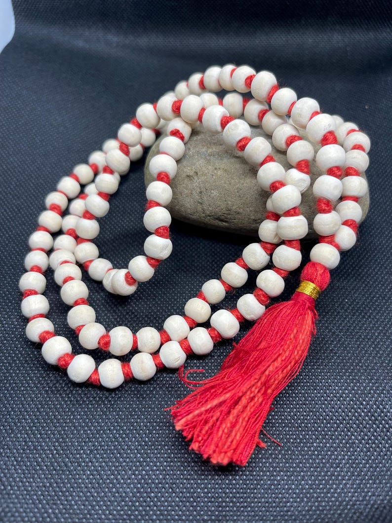 May include: A strand of round, off-white wooden beads strung on a vibrant red cord. The necklace features a prominent red tassel at the end, secured with a small gold-coloured metallic band, creating a continuous loop of beads with a knotted design.