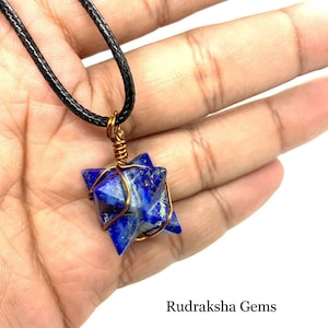May include: A blue lapis lazuli star pendant wrapped in copper wire, hanging from a black cord necklace. The pendant is held in a hand, with the text "Rudraksha Gems" visible below.