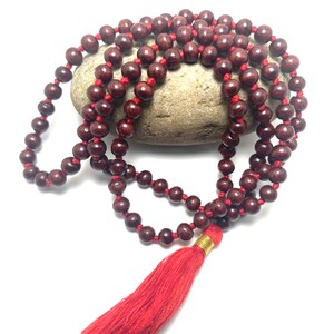 May include: A red wooden mala bead necklace with a red tassel. The beads are strung on a red cord and the tassel is attached to a gold bead.