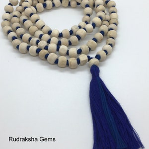May include: A string of light beige wooden beads separated by knots on a vibrant blue cord, ending in a long blue tassel. The text Rudraksha Gems is printed in the lower left corner. The beads are arranged in a circular pattern on a white surface.