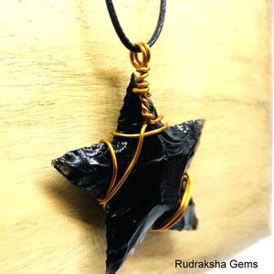 May include: A black obsidian star pendant wrapped in copper wire, hanging from a black cord necklace. The pendant is on a wooden surface. Rudraksha Gems is printed below the pendant.