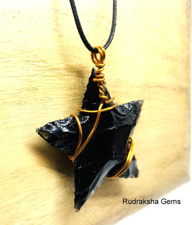 May include: A black obsidian star pendant wrapped in copper wire, hanging from a black cord necklace. The pendant is on a wooden surface. Rudraksha Gems is printed below the pendant.