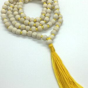 May include: A string of light-coloured, round wooden beads threaded on a bright yellow cord, arranged in a circular pattern. The strand features a prominent yellow tassel at the end. The text "Rudraksha Gems" is printed in the bottom left corner.