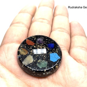 Puede incluir: Una piedra de curación de chakras redonda, negra, a base de resina, con destellos dorados y siete piedras de diferentes colores incrustadas. Rudraksha Gems está impreso en blanco en la parte superior de la imagen.