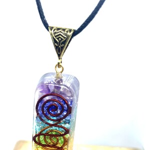 May include: A rainbow chakra pendant necklace with a gold-tone metal clasp. The pendant is made of clear resin with embedded crystals and a copper wire spiral design. The necklace is on a black cord.