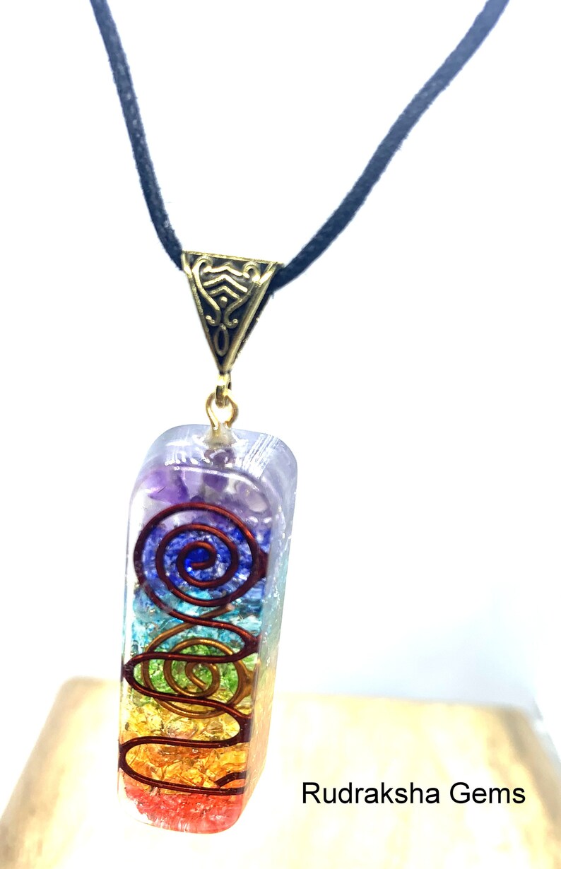 May include: A rainbow chakra pendant necklace with a gold-tone metal clasp. The pendant is made of clear resin with embedded crystals and a copper wire spiral design. The necklace is on a black cord.