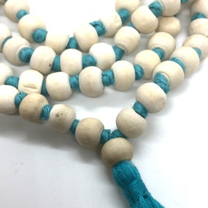 May include: A string of light beige, smooth, spherical wooden beads separated by knots of vibrant turquoise thread. The necklace features a matching turquoise tassel at the end. The text Rudraksha Gems is printed in black in the bottom left corner.