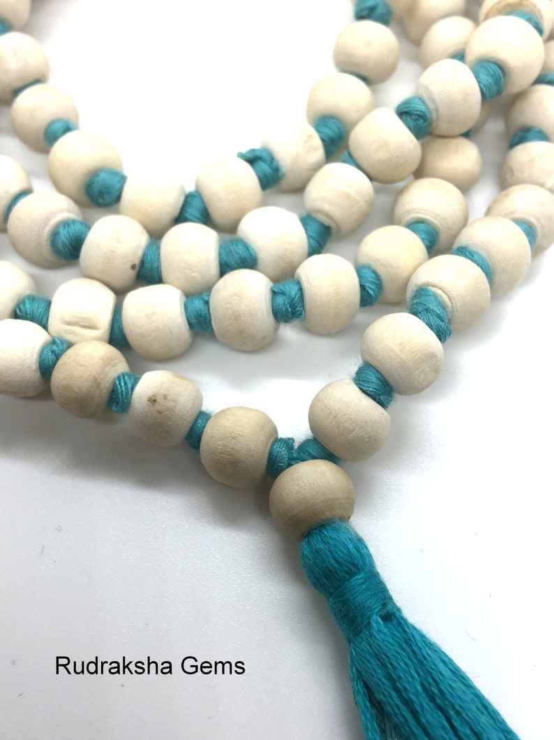 May include: A string of light beige, smooth, spherical wooden beads separated by knots of vibrant turquoise thread. The necklace features a matching turquoise tassel at the end. The text Rudraksha Gems is printed in black in the bottom left corner.