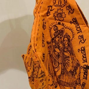 May include: Orange fabric pouch with a detailed illustration of Hindu deities, featuring Krishna and Radha. The design includes floral motifs and script in a dark brown color. The pouch has a loop for hanging.