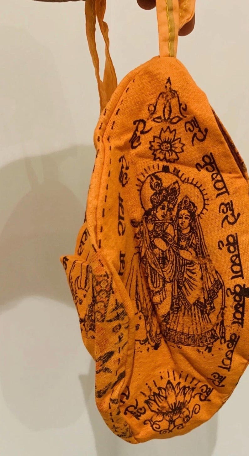 May include: Orange fabric pouch with a detailed illustration of Hindu deities, featuring Krishna and Radha. The design includes floral motifs and script in a dark brown color. The pouch has a loop for hanging.