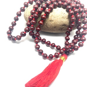 May include: A maroon wooden mala bead necklace with a red tassel. The mala beads are strung on a red cord.