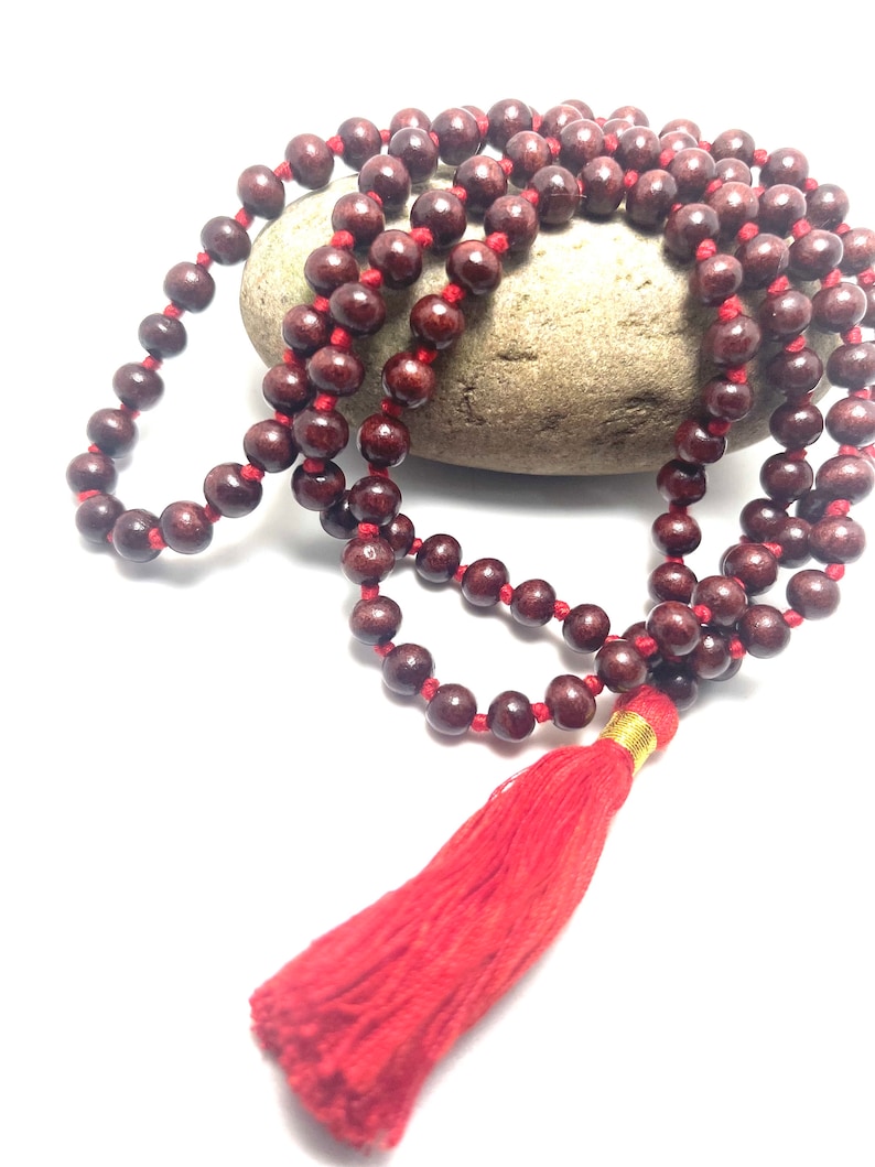 May include: A maroon wooden mala bead necklace with a red tassel. The mala beads are strung on a red cord.