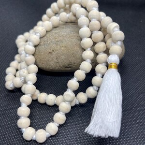 May include: A strand of light-coloured, natural wooden beads strung on a white cord, featuring a white fabric tassel at one end secured with a thin gold-coloured metallic band. The beads are uniform in size and shape, arranged in a circular loop.