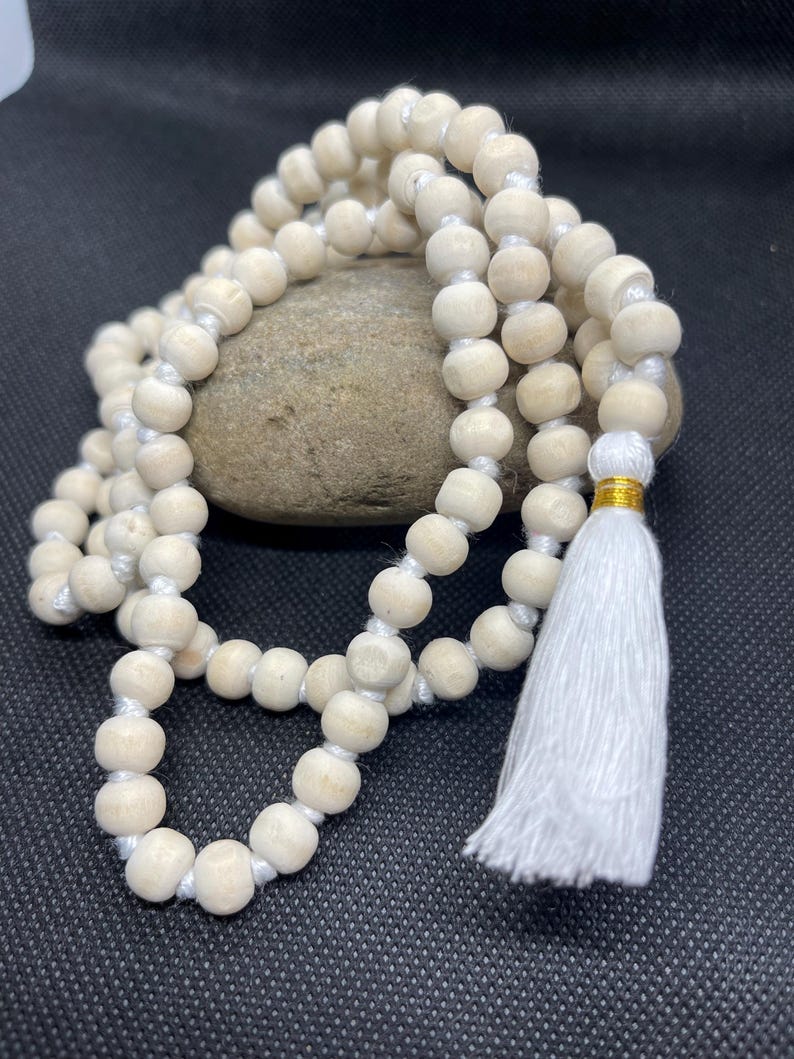 May include: A strand of light-coloured, natural wooden beads strung on a white cord, featuring a white fabric tassel at one end secured with a thin gold-coloured metallic band. The beads are uniform in size and shape, arranged in a circular loop.