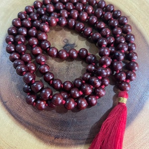 May include: A dark red wooden mala bead necklace with a red tassel. The beads are round and have a smooth finish. The tassel is made of a soft, silky material.
