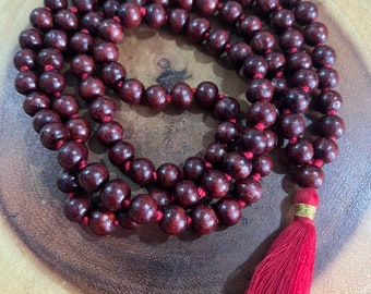 May include: A dark red wooden mala bead necklace with a red tassel. The beads are round and have a smooth finish. The tassel is made of a soft, silky material.