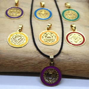 Może przedstawiać: Siedem złotych zawieszek z różnokolorowymi wkładkami emaliowanymi, każda z innym symbolem czakry. Zawieszki znajdują się na czarnym sznurkowym naszyjniku.