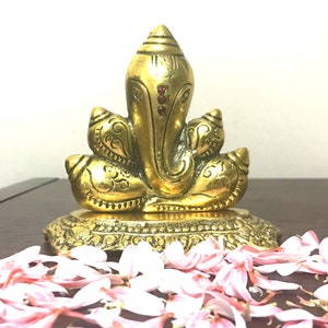 Puede incluir: Una estatua dorada de lat&oacute;n del dios hind&uacute; Ganesha, sentado sobre una base decorativa. La estatua presenta detalles intrincados y est&aacute; adornada con peque&ntilde;os acentos rojos. P&eacute;talos de flores rosas y blancas est&aacute;n esparcidos alrededor de la base.