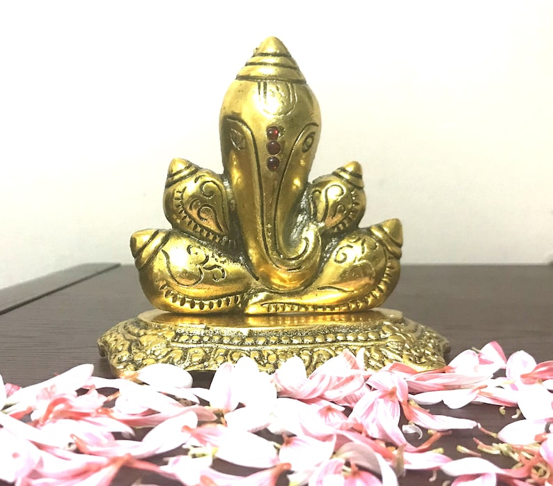 Puede incluir: Una estatua dorada de lat&oacute;n del dios hind&uacute; Ganesha, sentado sobre una base decorativa. La estatua presenta detalles intrincados y est&aacute; adornada con peque&ntilde;os acentos rojos. P&eacute;talos de flores rosas y blancas est&aacute;n esparcidos alrededor de la base.