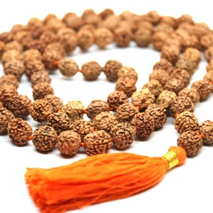 Peut inclure: Un collier de perles mala brun avec un pompon en fil orange. Les perles sont faites de matériaux naturels et ont une texture rugueuse.