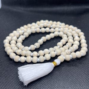May include: A strand of light-coloured, round wooden beads strung on a white cord. The necklace features a white fabric tassel at the bottom, secured with a small band of metallic gold thread. The beads are arranged in a circular, coiled pattern.