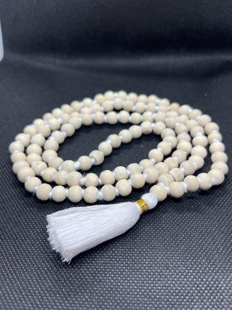 May include: A strand of light-coloured, round wooden beads strung on a white cord. The necklace features a white fabric tassel at the bottom, secured with a small band of metallic gold thread. The beads are arranged in a circular, coiled pattern.