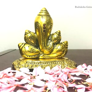 Puede incluir: Una estatua dorada de Ganesha, la deidad hind&uacute;, con detalles intrincados y acentos rojos. La estatua est&aacute; sobre una base decorativa, rodeada de p&eacute;talos de flores rosas. El texto "Rudraksha Gems" es visible.
