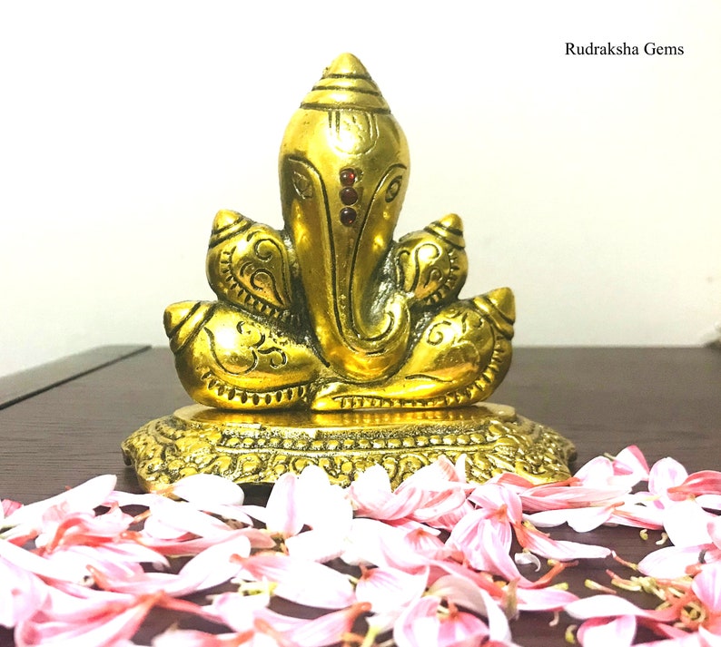 Puede incluir: Una estatua dorada de Ganesha, la deidad hind&uacute;, con detalles intrincados y acentos rojos. La estatua est&aacute; sobre una base decorativa, rodeada de p&eacute;talos de flores rosas. El texto "Rudraksha Gems" es visible.